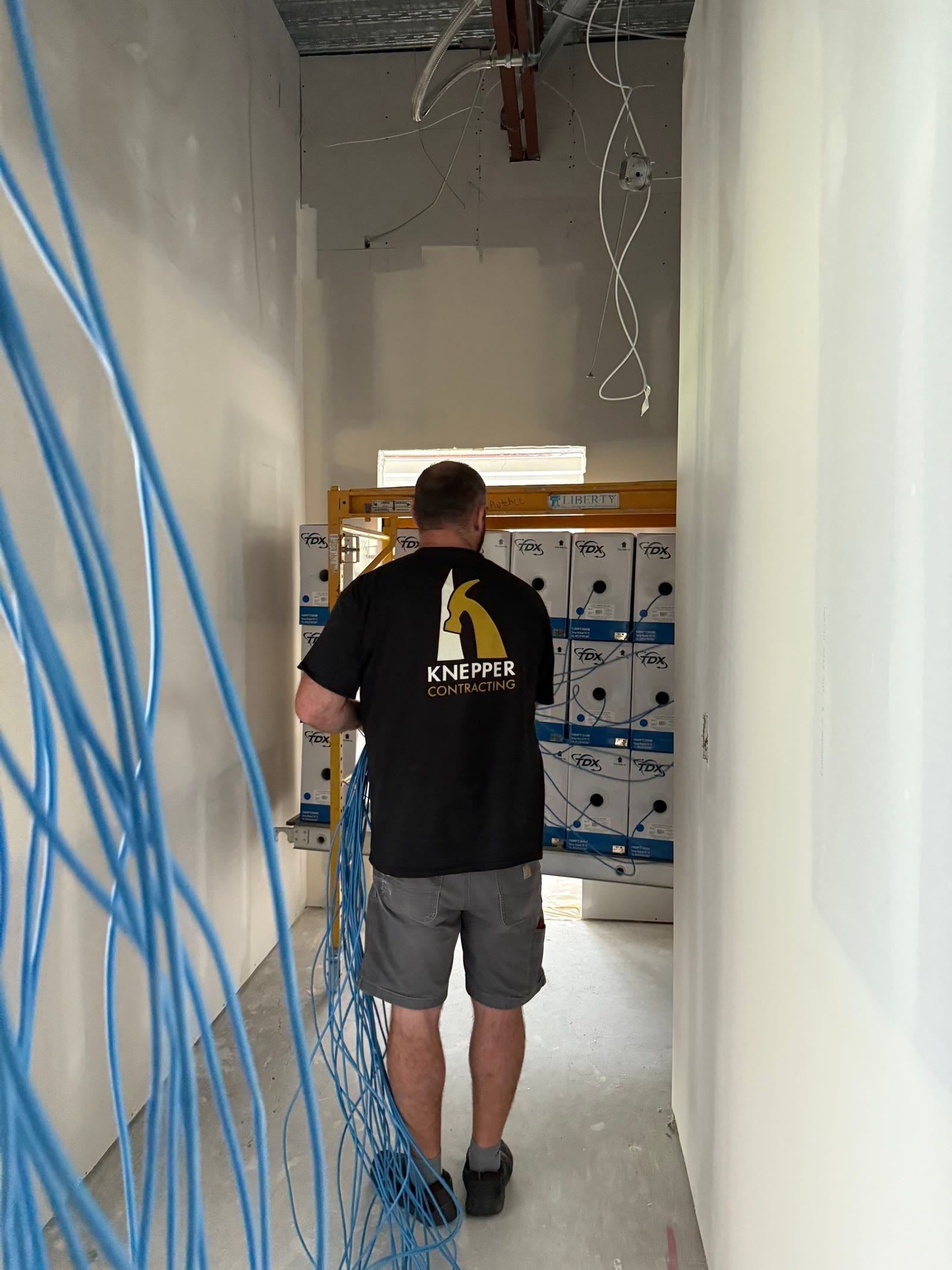 A Knepper Contracting technician, branded shirt visible, pulling cable off spools in an unfinished hallway.