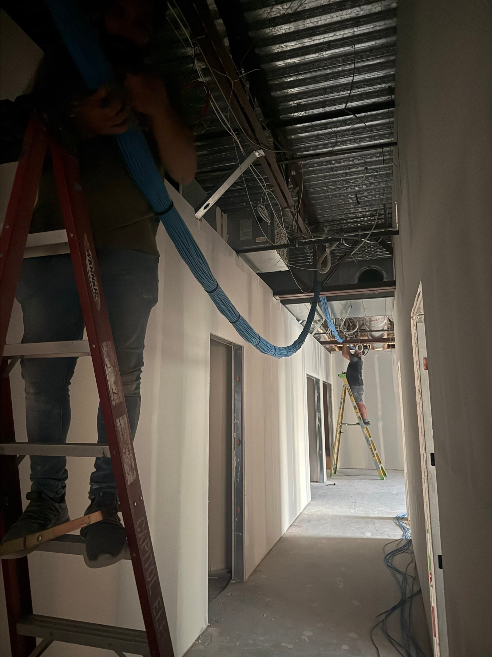 Two Knepper technicians on ladders running a main cable bundle overhead in an under-construction hallway.