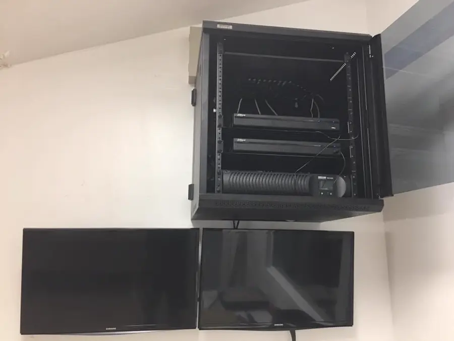 A small locking CCTV rack with a patch panel, two NVR/DVR units and a UPS above two mounted monitors.
