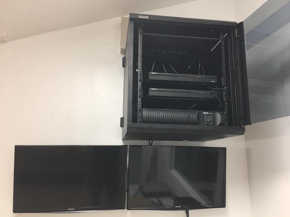A small locking CCTV rack with patch panel, two recorders and a UPS above two mounted monitors