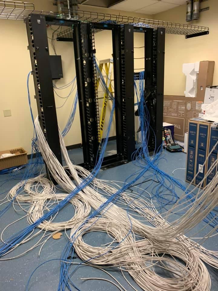 Wiring closet mid-job with two open racks, coiled cable on a raised floor, and displays staged nearby awaiting mounting.
