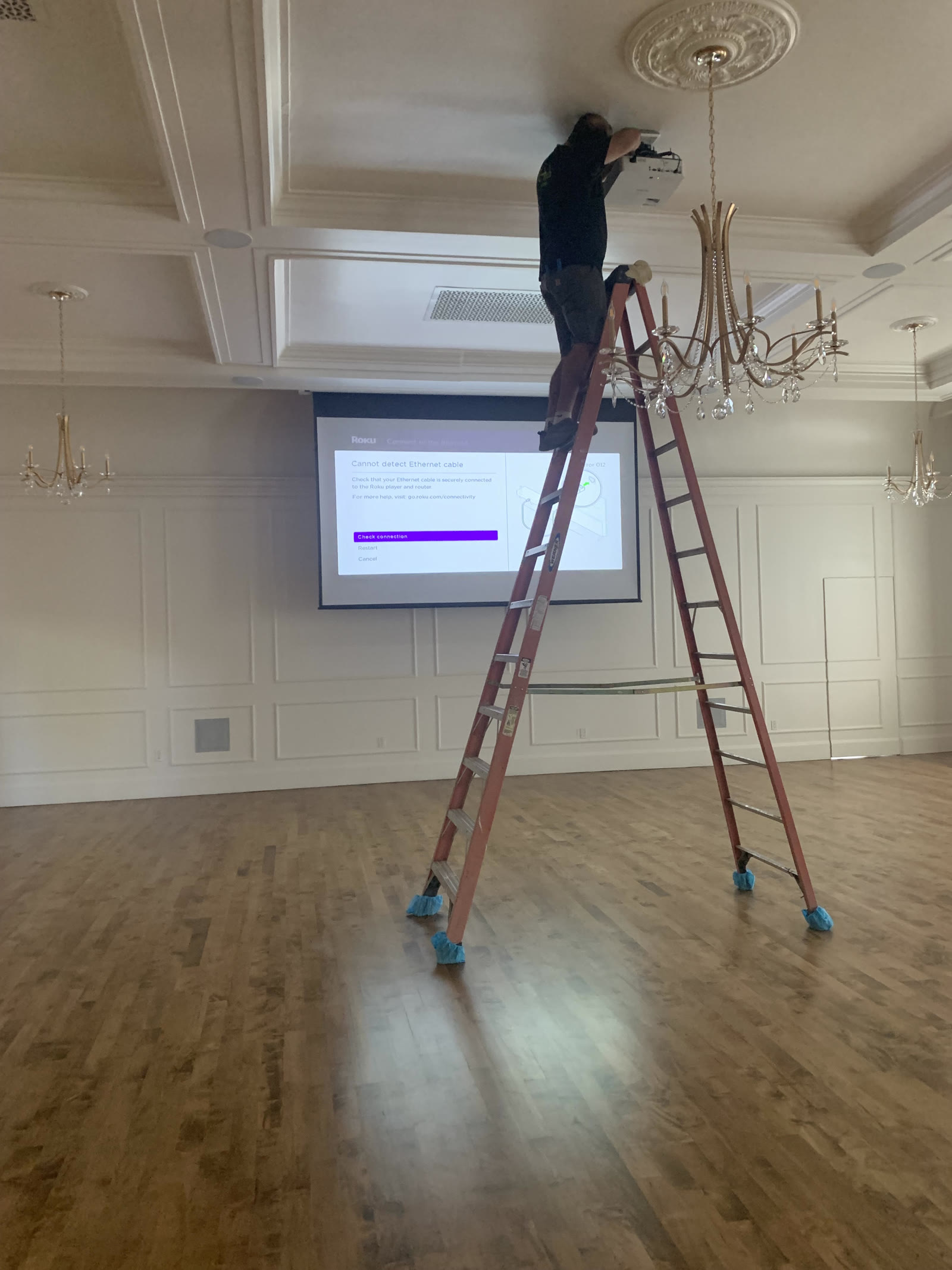 Technician on a ladder servicing a ceiling-mounted projector in a large event space.