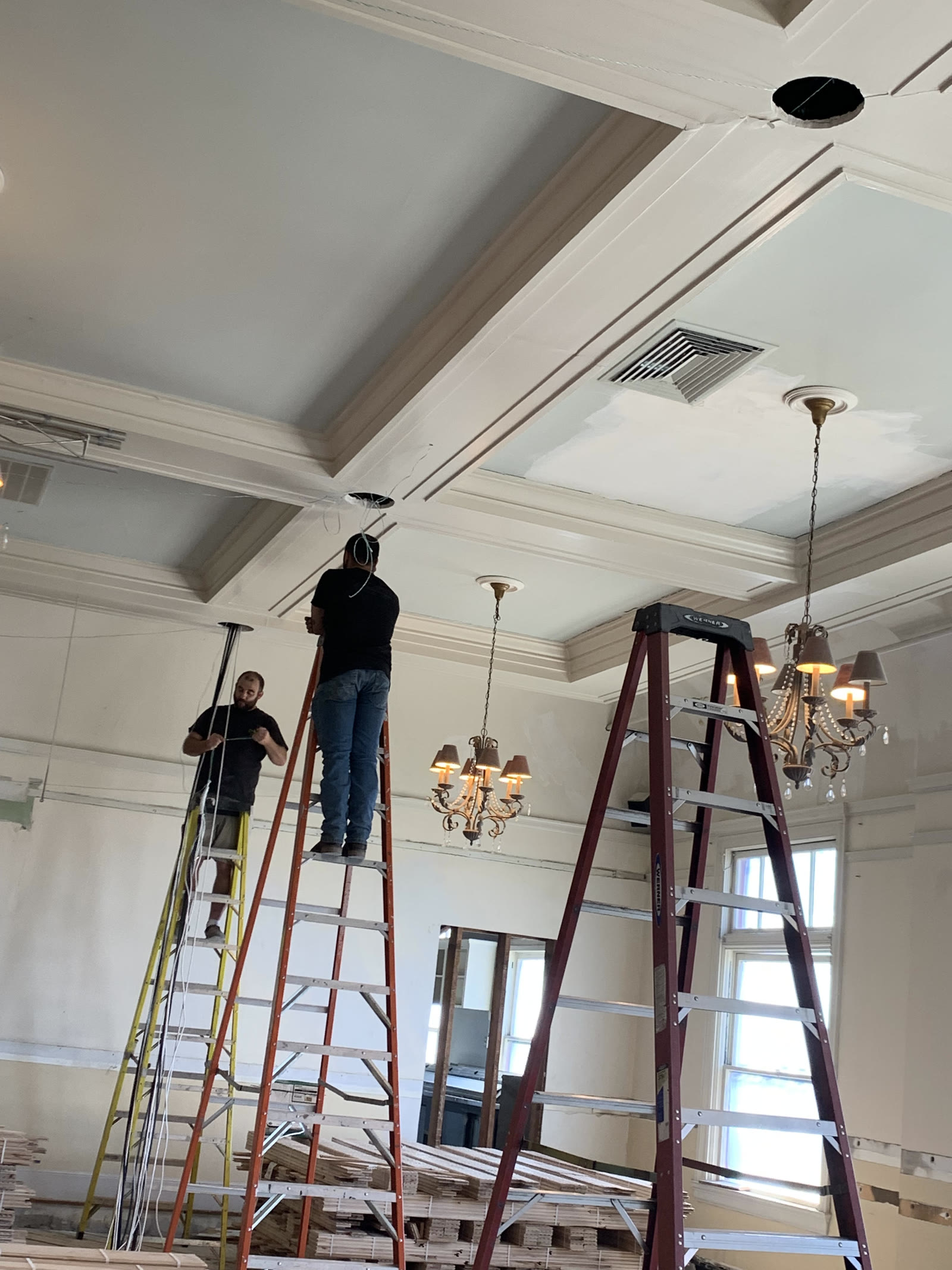 Two crew members on ladders wiring the ceiling of a mid-renovation ballroom